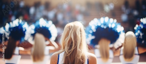 A group of cheerleaders, clad in matching uniforms with blue pompons, stand in front of a mirror, their reflections bouncing back. The basketball court provides a backdrop to their synchronized photo