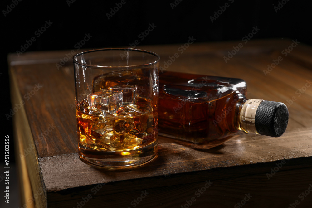 Whiskey with ice cubes in glass and bottle on wooden crate against black background, closeup