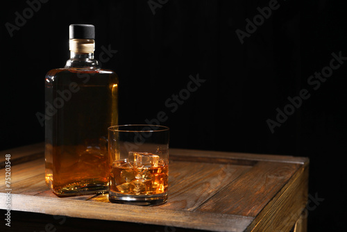 Whiskey with ice cubes in glass and bottle on wooden crate against black background, space for text