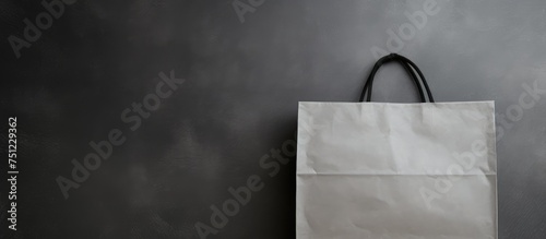 A white paper bag with a black handle is hanging on a grey wall. The bag appears to be empty and is neatly placed against the wall.