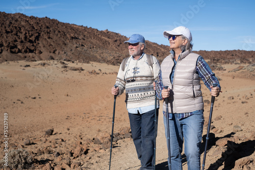 Active elderly retired couple with backpack walking outdoors with help of poles enjoying nature, freedom and free time. Arid terrain at the base of Teide volcano on the island of Tenerife