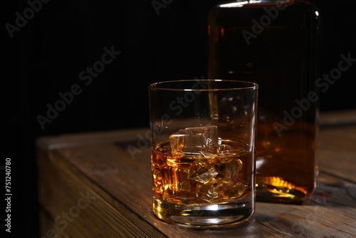 Whiskey with ice cubes in glass and bottle on wooden crate against black background, closeup. Space for text