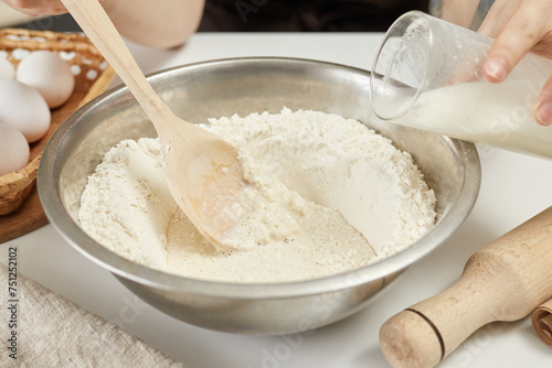 Unrecognizable woman hands adding milk and other bakery ingredients to flour preparing dough for baking bread in home kitchen
