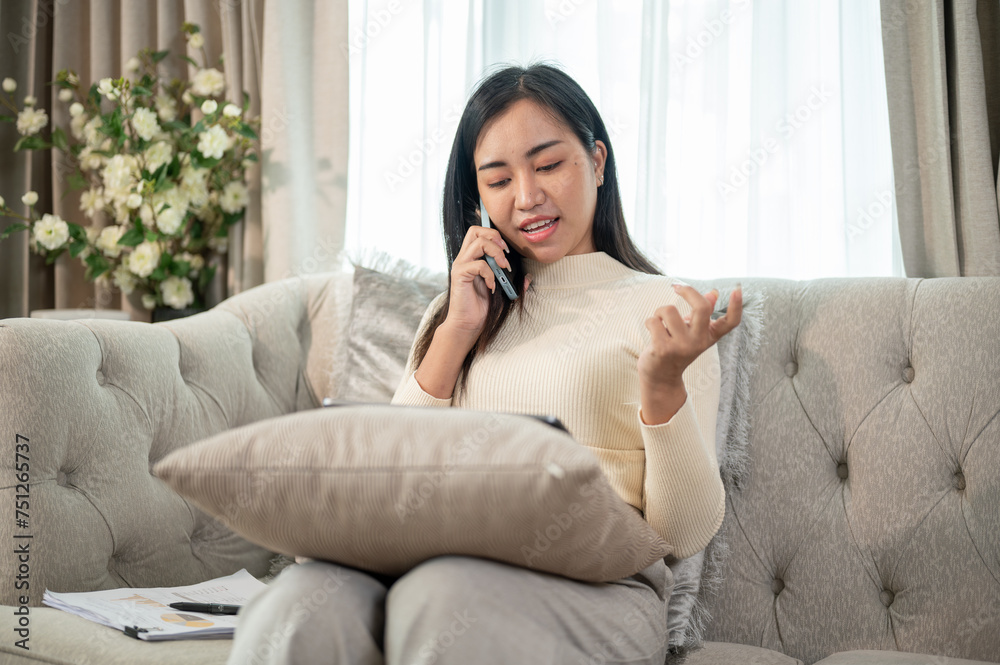 A positive Asian businesswoman is working remotely at home, conducting business over the phone. A positive Asian businesswoman is working remotely at home, conducting business over the phone.