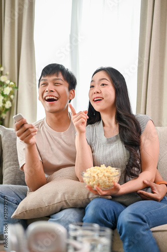 Happy young Asian couple eating popcorn while enjoying watching television on a sofa.