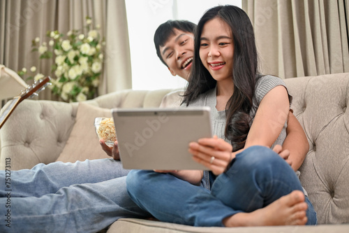Happy Asian couple watching a movie on a digital tablet while relaxing on a sofa at home together.