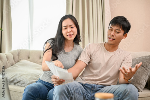 A resentful, angry Asian wife and husband are arguing about family household invoices on sofa.