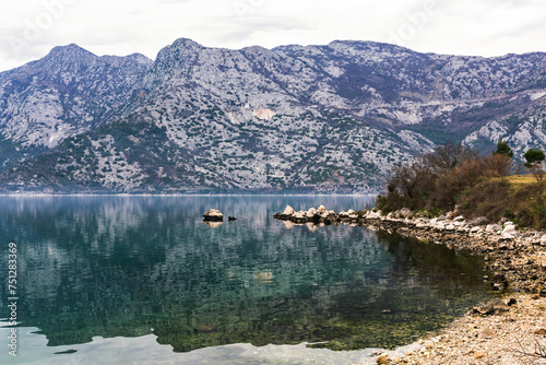 Reflective waters of Kotor Bay reveal rugged mountains, with scattered rocks lining the shore in Montenegro's serene landscape photo