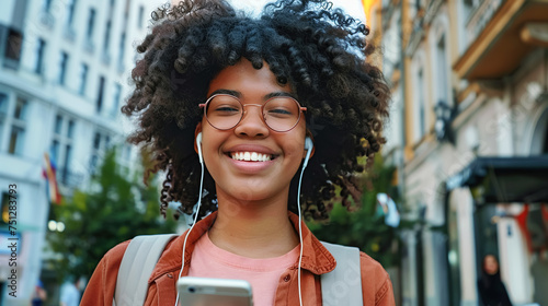 Happy young African woman wearing headphones and holding phone, looking at camera and smiling, walking along a busy city street. Modern communication, lifestyle and travel © ximich_natali