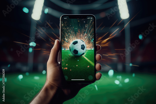 Hand holding a smartphone displaying a soccer ball with spark effects at a stadium. photo