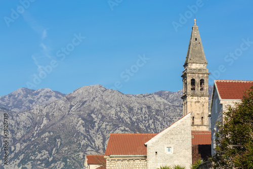 Bell tower rises against a mountain backdrop in Perast, Montenegro; a red-tiled roof and stone walls highlight historical architecture typical of the Adriatic