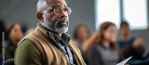 An African American man stands in front of a group of people, listening attentively and taking notes during an adult education class. He appears focused and engaged in the lecture.