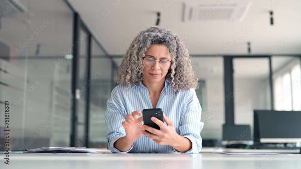 Busy mature professional businesswoman executive using smartphone cell mobile apps on cellphone working sitting at desk. Middle aged business woman holding phone using cellphone in office.