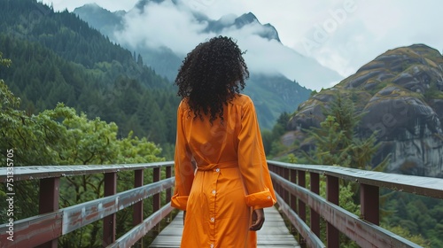 Full length of optimistic black woman in orange button-down dress strolling across a footbridge and staring off into the distance against a backdrop of greenery and mountains