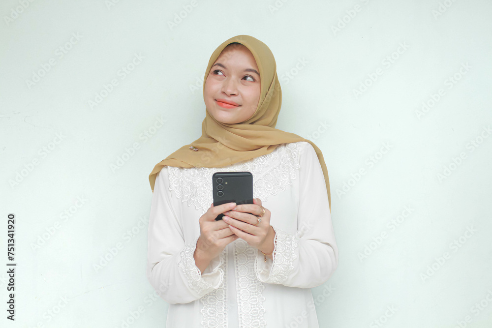 Young Asian Muslim woman holding mobile phone with happy expression with thinking something. Isolated in gray background. Young Asian Muslim woman holding mobile phone with happy expression with thinking something. Isolated in gray background.