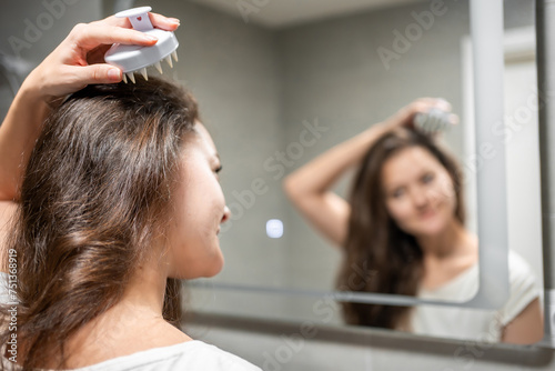 Young woman doing self hair scalp massage with scalp massager or hair brush for hair growth stimulating at home. Reflected view of the mirror