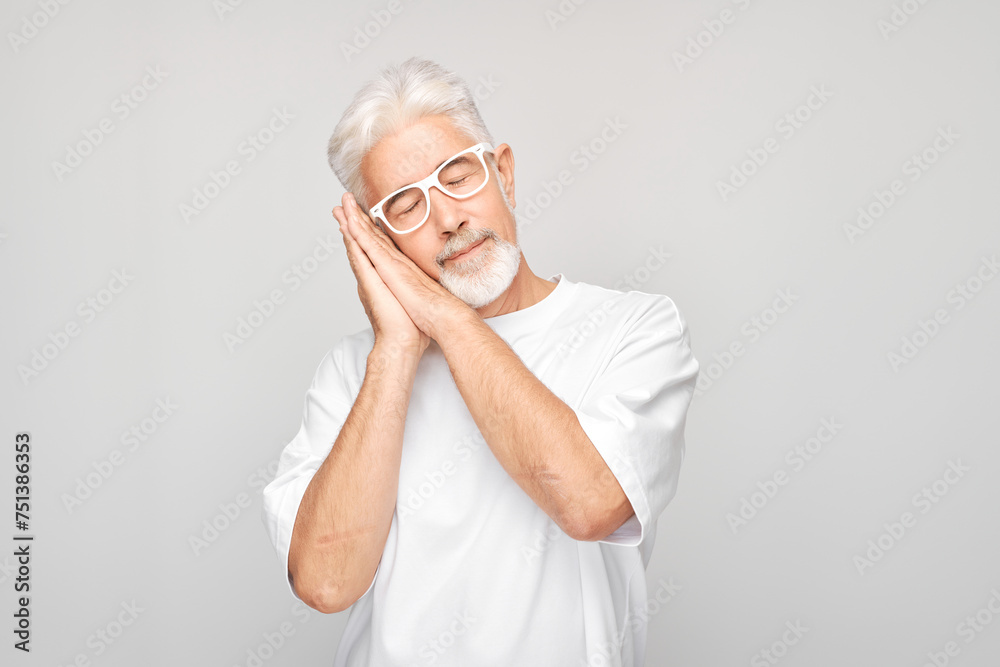 Senior man with glasses pretending to sleep, standing against a gray background.