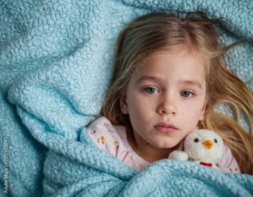 A little girl is laying in bed with a toy. She is looking at the camera with a sad expression