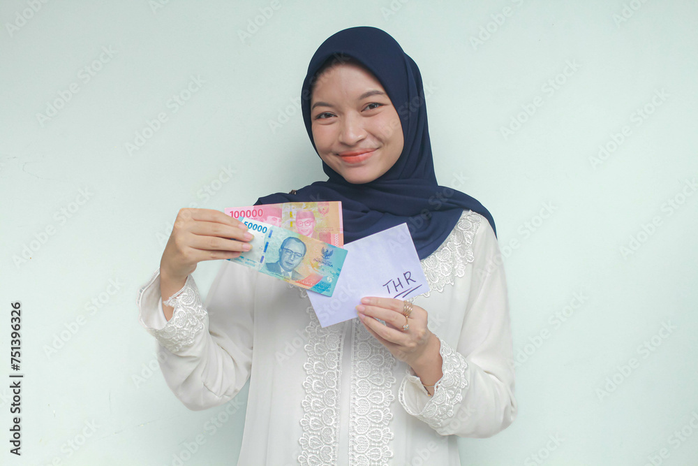 A young Asian muslim woman holding an envelope and one hundred and fifty thousand rupiah in cash with an expression of happiness. THR (Tunjangan Hari Raya) or Religious Holiday Allowance Concept. A young Asian muslim woman holding an envelope and one hundred and fifty thousand rupiah in cash with an expression of happiness. THR (Tunjangan Hari Raya) or Religious Holiday Allowance Concept.