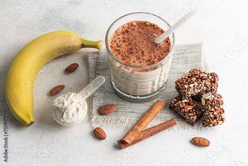 Glass with protein drink, healthy milkshake smoothie on white table with bananas, protein powder in measuring spoon, protein bar, almond nuts and cinnamon sticks