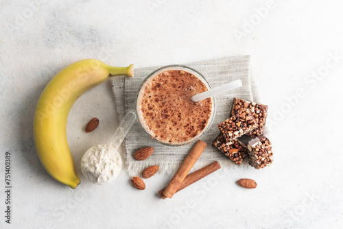Glass with protein drink, healthy milkshake smoothie on white table with bananas, protein powder in measuring spoon, protein bar, almond nuts and cinnamon sticks, top view