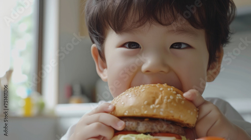 A boy is sitting in the kitchen  holding a big juicy burger in his hands and smiling happily.