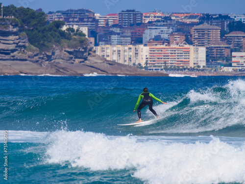 A surfer in a wetsuit rides a wave, with the city of Santander as a backdrop.