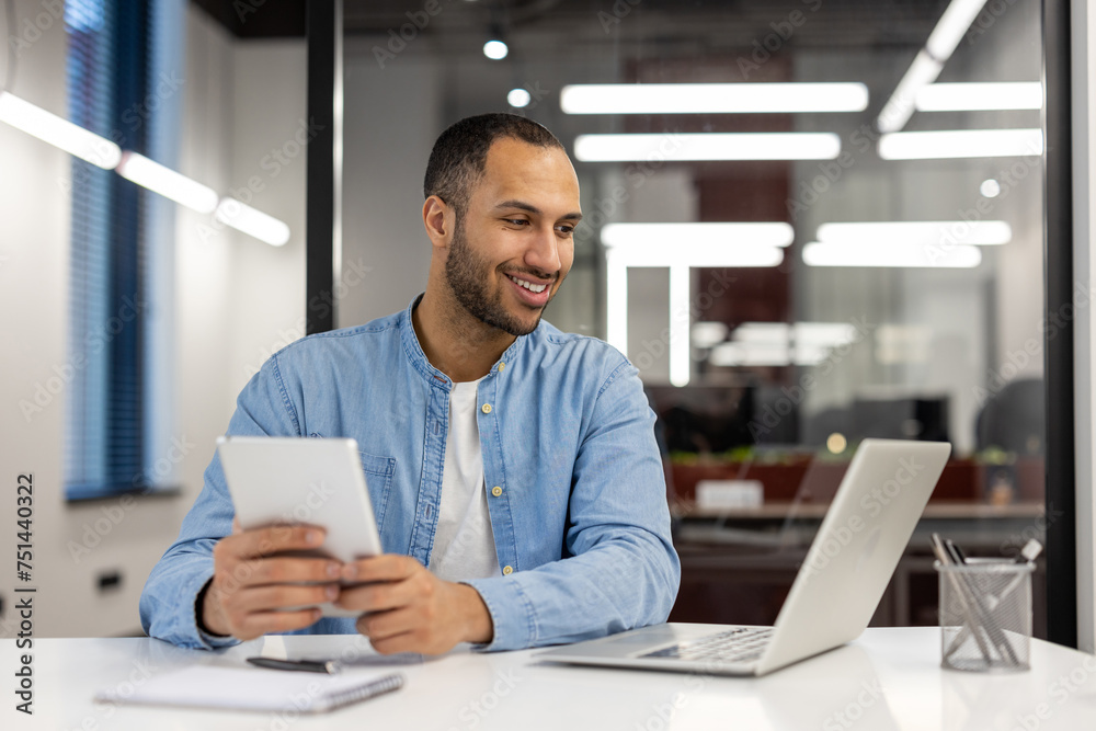 A young Hispanic man is talking on a video call on a laptop, sitting at a desk in the office, looking at the screen with a smile and holding a tablet in his hands A young Hispanic man is talking on a video call on a laptop, sitting at a desk in the office, looking at the screen with a smile and holding a tablet in his hands