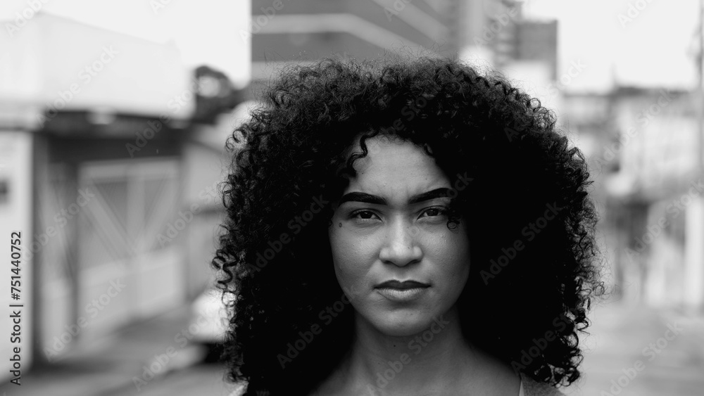Intense Gaze of African American Young Woman in Street, Black and White Close-Up Face in Urban Environment. A Brazilian adult girl in 20s with curly hair with stern confident expression