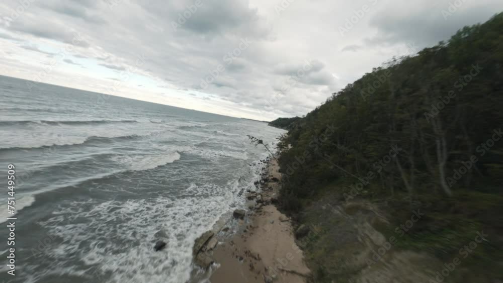 FPV drone flight above picturesque view of foamy sea waves rolling sandy beach with rocks near cliff under cloudy gloomy sky