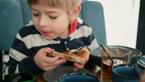 Cute healthy preschool kid boy eats a piece of cheese pizza for lunch. Charming child biting off big slice of fresh made pizza at the table in a restaurant indoor. Funny little toddler. Unhealthy food photo
