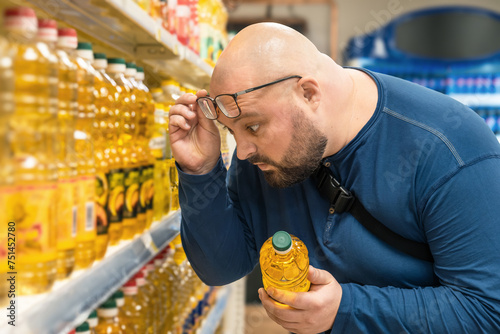 Shocked overweight man raising his glasses to his forehead and looking on price of vegetable oil in supermarket. Amazed male by food inflation and rising prices on sunflower oil in grocery store