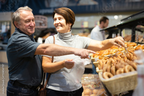 Elderly married couple chooses hot fresh buns and bread at supermarket showcase