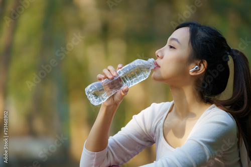 Asian woman drinking water from a bottle after running with a public park background, outdoor workout concept, drinking clear mineral water after jogging.