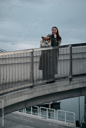 A young woman poses with a dog standing on a beautiful architectural bridge in the center of Belgrade on the embankment. Female owner with red welsh corgi Pembroke, urban lifestyle. © Ekaterina