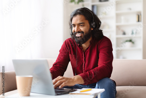 Cheerful indian man working from home, using headset and laptop