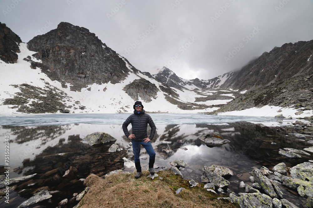 Happy active cheerful tourist in mountain sunglasses and a down jacket, joyful smiles expressively rejoices against the backdrop of a mountain lake high in the mountains
