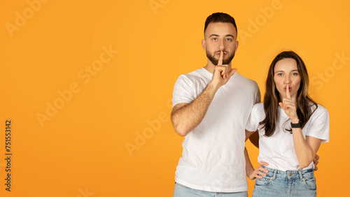 Quietly confident man and woman in white shirts placing fingers on lips in a 'shush' gesture