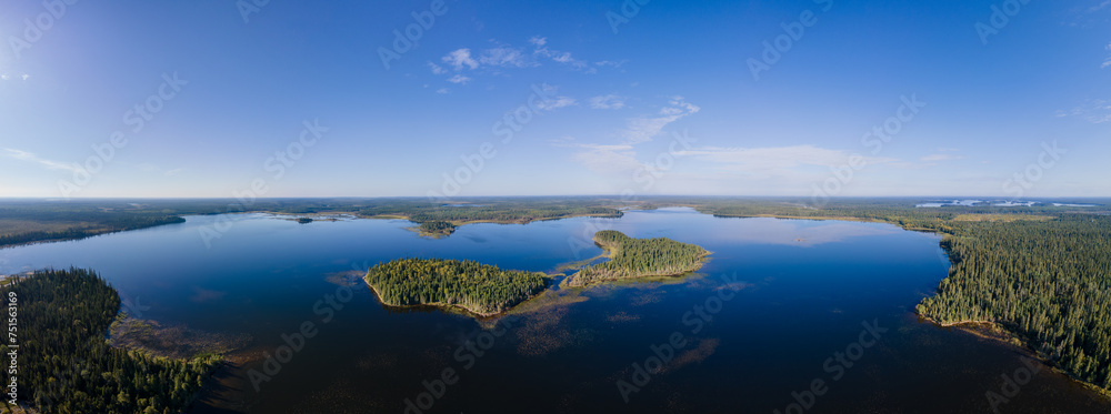 Aerial panoramic view of a northern wilderness lake with small islands. The water is calm and reflecting the blue sky.  The surrounding forest is a mix of spruce, pine, and aspen trees.
