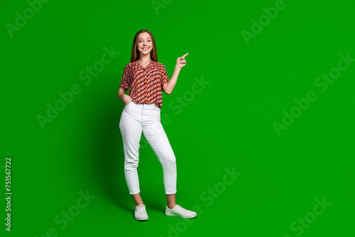 Full size photo of positive teen girl wear print blouse white trousers indicating at offer empty space isolated on green color background