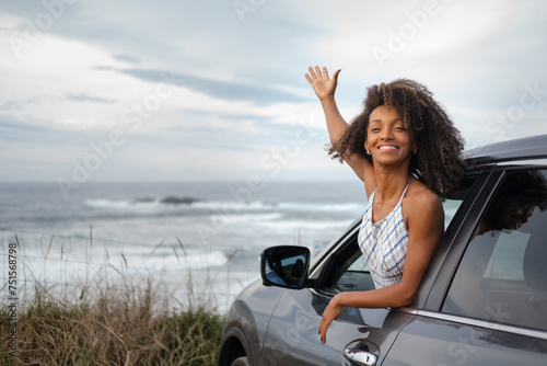 Blissful young black woman leaning out of the window of her car with the sea and sky in the background. Female driver enjoying the freedom of a getaway.