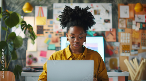 A young black woman in a creative office space, deeply focused on her laptop screen, with sketches and mood boards in the background, business technology, with copy space