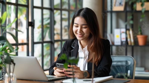 Young smiling asian businesswoman using and looking at mobile phone during working on laptop computer at modern office