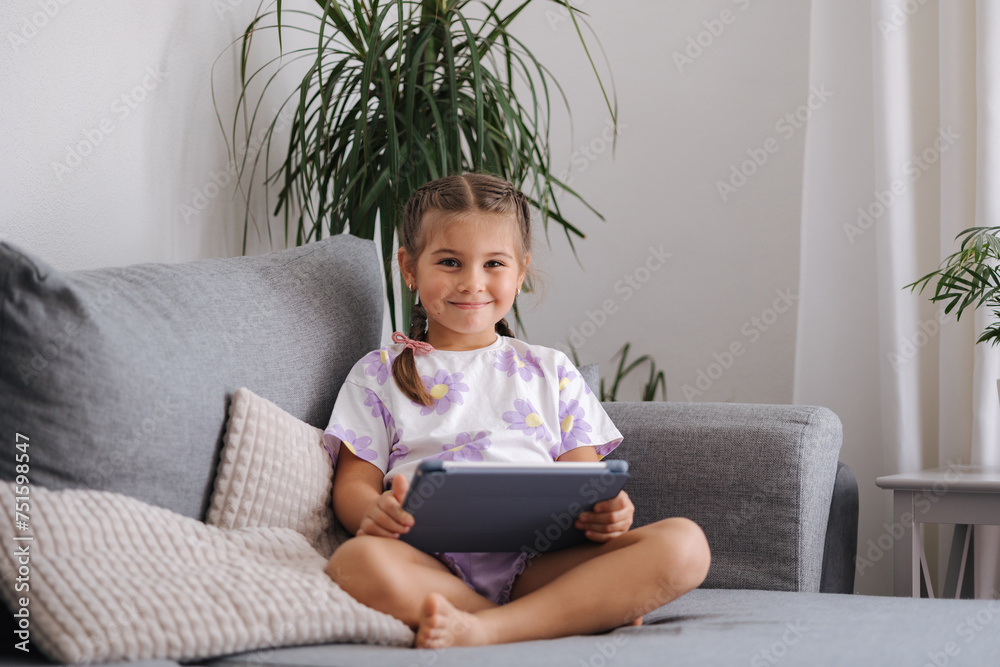 Adorable little girl sits at home on sofa and play games on tablet. Home mood