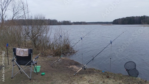 A folding fishing chair, a plastic bucket with bait, a cage and feeder rods for bream fishing mounted on stands on the ground next to bushes on the river bank. Flood. On the far bank is a forest