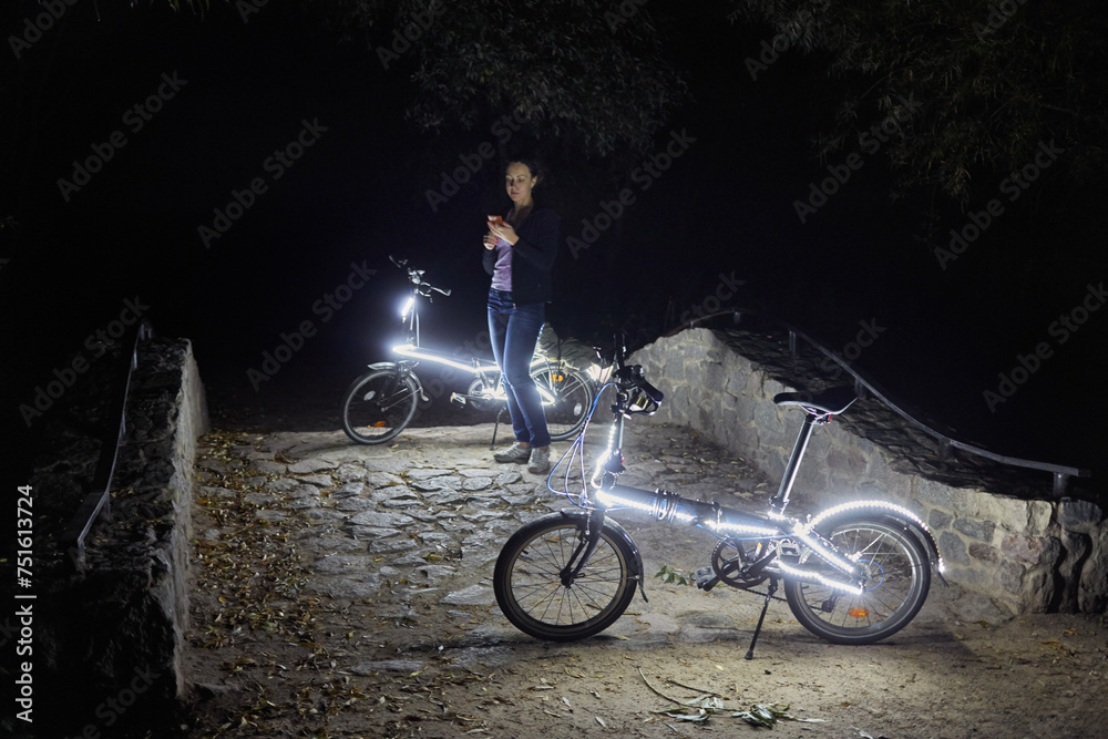 Obraz premium Woman with cell phone stands at night park near two bicycles decorated with luminous diodes