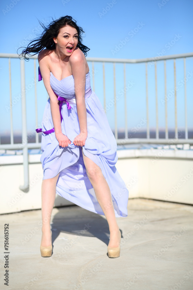 Pretty girl in dress stands on roof of tall building and shouts on wind at sunny day