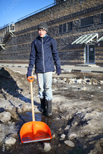 Bild auf Leinwand Boy in black stands with shovel among melted snow at sunny spring day