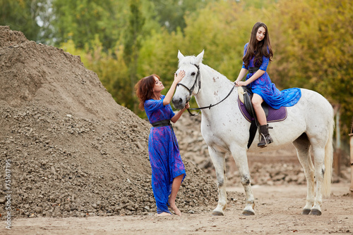 Woman holds by bridle white horse on which her daughter sits in park near ground pile