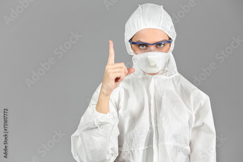 Half-growth portrait of woman in white protective suit of synthetic paper and respiritory half mask, looking at us, finger up, on gray background photo